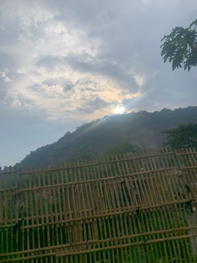 A wooden fence made of horizontal and vertical slats, likely bamboo, is in the foreground of the image. The fence is situated in front of a hillside covered in foliage. The sun is shining through the clouds in the sky, casting beams of light onto the scene. A portion of the hillside appears to have a visible scar, possibly from a landslide. The coordinates provided indicate the scene is located in a rural area of the city of Lagadar, Indonesia. No people or other subjects are visible in the image.