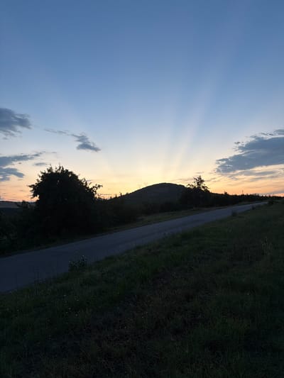 This is a landscape photograph taken in Shatrovo, Bulgaria, depicting a rural scene during sunset. The image is oriented vertically, with the sky occupying the left side and the ground and a road occupying the right.
The sky is a prominent feature, transitioning from a pale blue at the top to warmer hues of orange and yellow near the horizon, indicating the time of day is either sunrise or sunset. Sun rays are visibly fanning out from behind a dark, silhouetted hill or mound. Clouds are scattered across the sky, some appearing wispy and others more substantial.
The ground on the right is covered in dark green grass, with a paved road running diagonally through it. The road is bordered by vegetation, including trees and bushes that are also silhouetted against the fading light. The overall impression is one of tranquility and natural beauty. There are no people or specific objects visible, making the scene feel serene and empty. The time of day, with its soft, warm light, evokes a sense of peace. The presence of the road suggests it might be a country road or pathway. The weather appears clear, with the sunset being the dominant atmospheric condition.