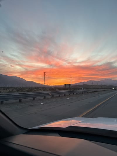 The image captures a vibrant sunset observed from inside a moving vehicle on a highway near Desert Hot Springs, United States. The sky displays a gradient of colors, transitioning from pale blue at the top to bright orange and red near the horizon. The clouds are streaked with these colors. The landscape features mountains in the distance, silhouetted against the fiery sky. Multiple wind turbines are visible in the mid-ground, along with telephone poles lining the highway. The highway itself is divided by a yellow line, with a barrier visible on the side of the road. The hood of the vehicle is partially visible in the foreground, reflecting the colors of the sunset. The scene suggests a journey at dusk, with the warm colors of the sky contrasting against the landscape.
