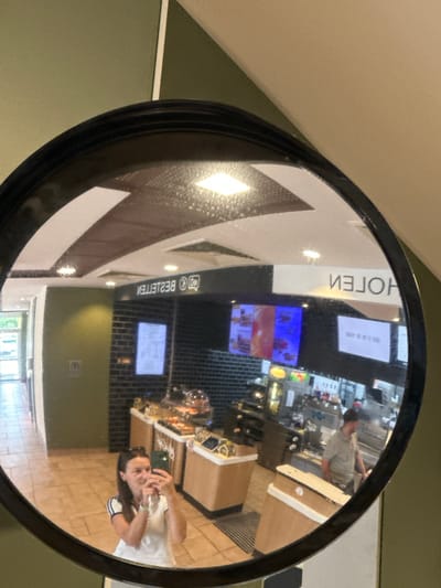 A round convex mirror displays the reflection of the interior of a building. Reflected in the mirror is part of a cafe or food court, showing a counter, display cases with food items, and people. One person, closer to the camera and presumably taking the picture, is holding a phone. The reflection suggests the room is brightly lit with various light fixtures. A sign in the reflection reads "HOTEL" (reversed) and "BESTELLEN" (reversed). The city is Ottersberg, Germany.