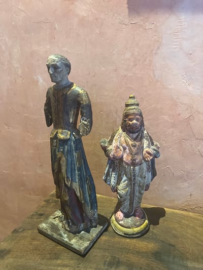 Two weathered, antique statues stand against a textured peach wall and a wooden surface. The lower statue, possibly male, has a weathered head and a dark, possibly blue, cloak. Gold detailing is visible. The upper statue, perhaps religious, features a crowned figure in lighter robes, with hands extended. Both figures display significant age and wear.