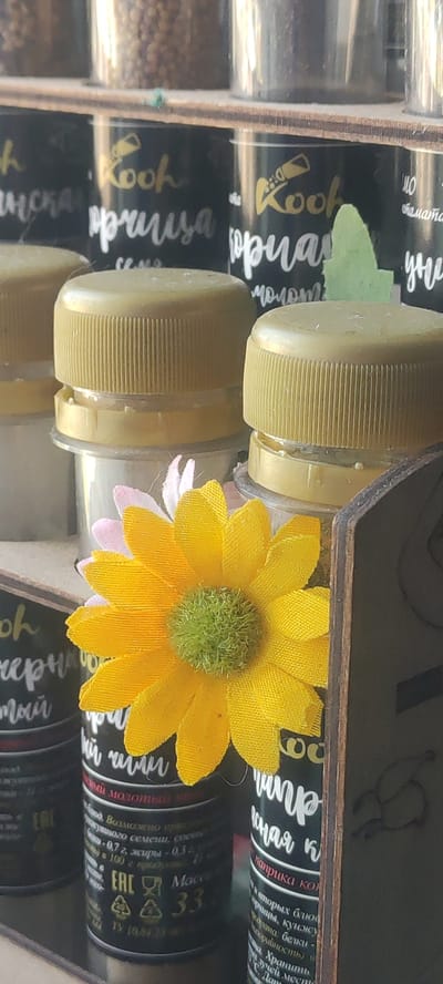 The image shows a close-up view of a shelf displaying several spice jars. The jars have black labels with white and gold lettering. Some of the labels are partially obscured by yellow bottle caps and a decorative artificial yellow and pink flower. The jars appear to contain ground spices or seeds. The overall scene is indoors, likely in a home or a small shop. The exact location in Ridder, Kazakhstan is not discernible from the image itself.