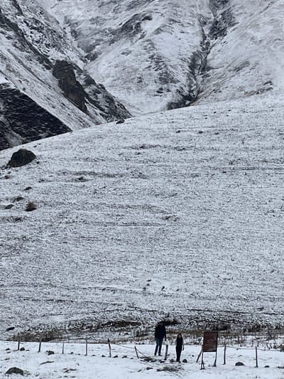 This image depicts a snow-dusted mountainous landscape in the vicinity of Juta, Georgia. In the foreground and midground, a gently sloping terrain is covered with a thin layer of white snow, revealing underlying darker ground and sparse vegetation. The background features steep, rugged mountains, partially covered in snow with exposed dark rock faces and visible erosion channels. In the lower right, two individuals are standing on the snow. The person on the left is taller, dressed in a dark winter jacket and dark pants, facing slightly right. To their right and slightly forward is a second, shorter individual in dark clothing, also facing right. Immediately to the right of these individuals, a dark rectangular sign on a post is visible, its text unreadable. A rudimentary barrier or pathway marker, consisting of short wooden posts connected by a rope or wire, runs horizontally across the lower foreground. The sky is overcast, providing diffuse lighting.