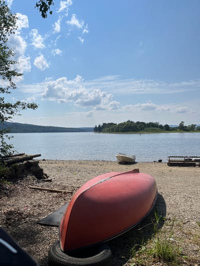 This is a photograph taken on a sunny day by a body of water, likely a lake, with a blue sky dotted with white clouds. In the foreground, a red overturned canoe rests on a tire and a black mat on a gravelly shore. To the right, a small white boat is partially visible on the shore, and further to the right is a wooden bench. Beyond the shore, the expansive lake stretches to a tree-covered shoreline with a hilly landscape in the background. The scene appears to be a recreational area near the town of Posudov in the Czech Republic, offering a peaceful outdoor environment.