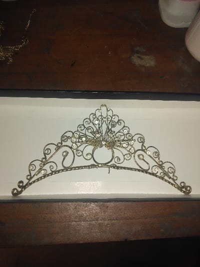 An ornate metal tiara is positioned centrally within a white-lined, black-rimmed rectangular container. The tiara is constructed from twisted, silver-toned wire forming an intricate openwork pattern, embellished with numerous small, clear, faceted stones. A distinctive fan-shaped motif with concentrated stones is centered at the top, while the sides display symmetrical scrollwork. The container rests on a dark brown wooden surface, which exhibits pronounced grain patterns, scratches, and several irregular dark and light stains. In the upper left quadrant of the image, a gold-colored chain is partially visible on the wooden surface. In the upper right quadrant, portions of two opaque objects, one white and one light pink, are visible. No individuals are present. This scene is located in the city of Gerasimovskoye, Kazakhstan.