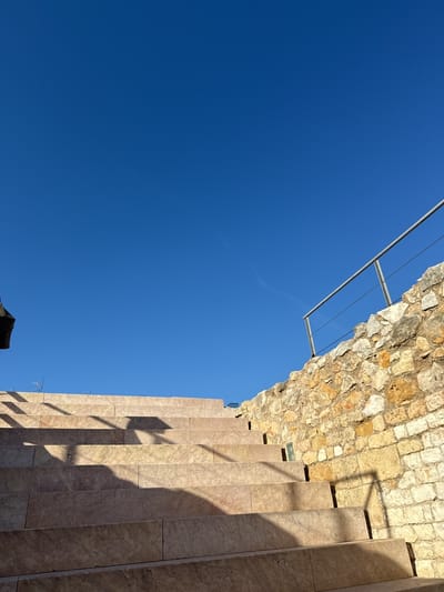 The image depicts a sunlit outdoor scene with a clear blue sky overhead. A set of stone steps rises diagonally across the lower half of the frame, casting shadows that add depth and texture. To the right of the steps, a rough stone wall is visible, topped with a simple metal railing. The architecture suggests a historical or public space.
Based on the architectural style and available information, it is likely that the location is in the city of Tarragona, Spain. However, definitive confirmation would require additional contextual information.