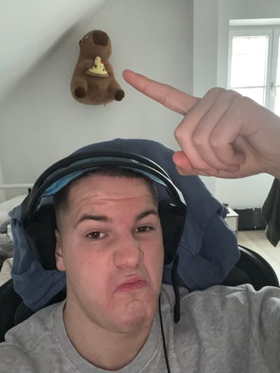 A selfie taken indoors, likely in a residential setting in Benningen, Germany, features a young adult male with short hair, light skin, and brown eyes. He's wearing a grey sweatshirt and a blue hoodie. He is wearing headphones around his neck. His face is positioned in the lower right of the frame. A hand with a pointing index finger is visible in the upper center of the image, directed towards a small, plush bear holding a slice of pizza. The bear is located on a white wall in the lower left of the frame. A window with a white frame is visible in the upper left corner, suggesting daylight. The room appears uncluttered.