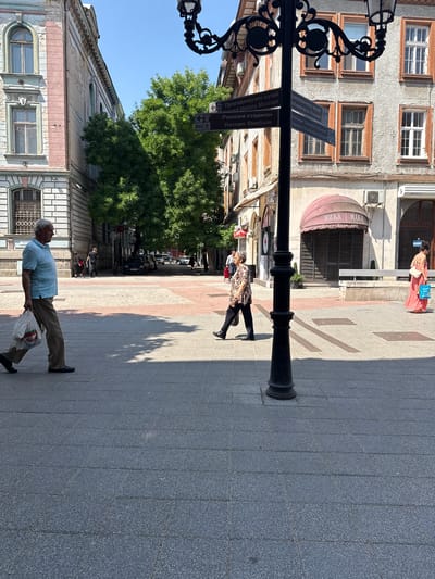 The image depicts a street scene in Plovdiv, Bulgaria, likely near the historic center. In the foreground, a man in a light blue polo shirt and khaki pants walks from left to right, carrying a white plastic bag. Behind him, another person walks in the same direction. In the middle ground, a woman in a patterned top and dark pants walks from right to left, and further back, a woman in a coral-colored dress walks from right to left carrying a shopping bag. A prominent black lamppost with ornate detailing stands in the center of the frame. Several street signs are attached to the lamppost, including those indicating "Natural History Museum," "Archaeological Museum," and "Roman Stadium." The background features several old buildings with architectural details and awnings. Trees with dense green foliage are visible between the buildings. The ground is paved with grey stone tiles. The scene is set on a sunny day with clear blue skies.