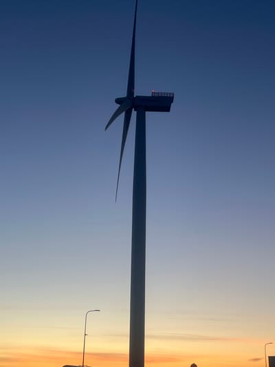A silhouetted wind turbine stands tall against a clear twilight sky. The sky transitions from a deep blue at the top to a soft yellow and orange gradient near the horizon, suggesting the end of a day. The wind turbine's nacelle has a red warning light visible. In the lower left of the frame, a streetlamp is partially visible. The scene is set in the vicinity of Nasva alevik, Estonia.