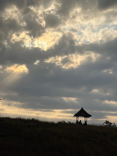 In Kubutambahan, Indonesia, at sunset, a silhouette of a traditional Balinese pavilion sits atop a grassy hill. The pavilion, with its thatched roof, shelters three figures seated inside, their forms indistinct against the dramatic sky. The sky is filled with heavy, dark clouds, through which rays of golden sunlight pierce, creating a dramatic and atmospheric effect. The foreground is a dark silhouette of dry grass covering the undulating terrain. To the left of the pavilion, two motorcycles are parked, also rendered in silhouette. The overall scene captures a moment of quiet contemplation at the end of the day, with the natural elements taking center stage.