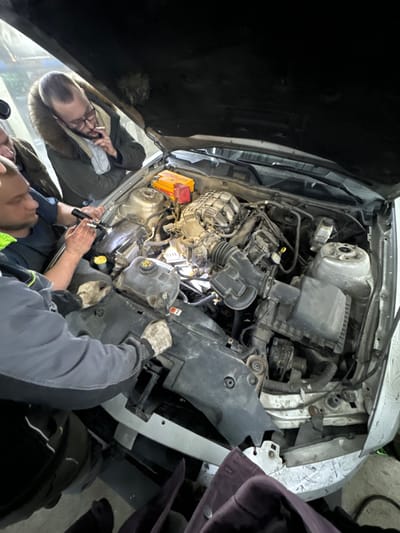 In Zhabkino, Russia, an elevated view captures an indoor scene within a garage or workshop. The primary subject is the open engine bay of a light-colored vehicle, revealing a somewhat soiled internal combustion engine and associated components. A red and yellow portable power or jump starter unit is visible on the passenger side of the engine bay.
Four adult males are engaged with the vehicle. In the center-left, a male with short dark hair and a beard, wearing a dark shirt with neon green trim, holds a black flashlight, directing its beam into the engine's deeper sections. Immediately to his right, another male, partially obscured, is observing. Above them, a male with a short dark beard, glasses, and wearing a dark, fur-hooded jacket over a light scarf, holds a lit cigarette between his fingers near his mouth as he looks down into the engine bay. In the lower-left foreground, a fourth male, identifiable by his gray work garment and beige work gloves, has both hands positioned on a black plastic shroud covering the front of the engine bay. The combined actions indicate a collaborative inspection or diagnostic process of the vehicle's engine. The environment is illuminated by artificial light sources.