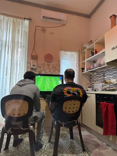 Two adult males are seated with their backs to the viewer in a residential room in Stornara, Italy, playing a soccer video game on a television. The individual on the left wears a grey hooded sweatshirt and grey sweatpants. The individual on the right has a bald head and wears a black t-shirt with a large, multi-colored graphic on the back; his feet are propped up. Both are seated on dark brown plastic chairs with light-colored, perforated backrests, tied with white ribbons.

The television screen displays a vibrant green soccer field with players. The room features light-colored walls, possibly a light pink or beige. On the left wall, above a window with sheer white curtains, an air conditioning unit is mounted, showing a blue digital display. Below the AC unit, exposed wiring runs to a white electrical meter box and a small, round white device, possibly a smoke detector or alarm. A pink heart-shaped wall clock is visible below these components.

To the right, a kitchen area is integrated into the space. It includes a white countertop with a stainless steel sink and faucet, a grey and white checkered tile backsplash, and a black oven below. A red towel hangs from the oven handle. Above the counter are open shelves displaying dishes and various household items, alongside a white upper cabinet. A patterned rug covers the floor.