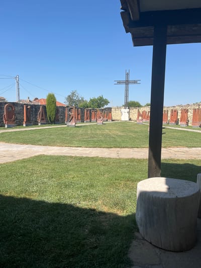 The image shows an outdoor courtyard in the city of Pliska, Bulgaria, with a prominent display of large, carved stone letters set against a low stone wall. The letters appear to be in the Cyrillic alphabet and are arranged in a semi-circular fashion. In the background, a large, modern metal cross stands against a clear blue sky, and two white statues of figures in robes are visible between the letters and the cross. The foreground is dominated by a well-maintained green lawn and paved pathways. A wooden overhang and a large wooden log, possibly a bench or table, are visible in the right foreground, casting shadows on the grass. The overall scene suggests a historical or cultural site, possibly a museum or memorial.
