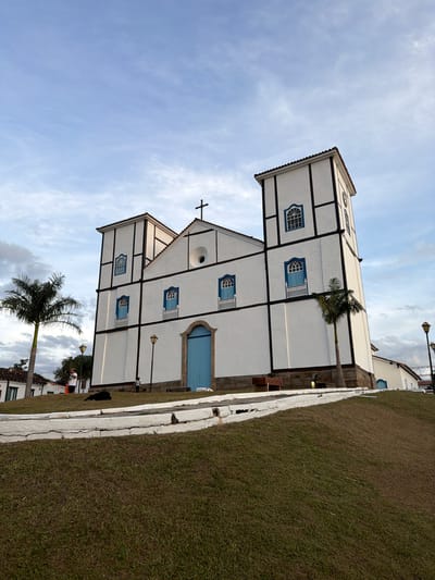 A street-level medium shot of the Igreja Matriz de Nossa Senhora do Rosário in Pirenópolis, Brazil, under a blue sky with wispy clouds. The church, a white, two-story building with a black frame and blue windows and doors, stands at an angle, with its front-facing side visible. A black cross sits atop the church. A palm tree partially obscures the view of the building. Adjacent to the church is a stretch of grass with a white curb. A few other buildings and some individuals are visible.