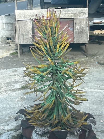 In Karama, Indonesia, a roughly constructed, artificial tree-like sculpture stands on a concrete surface. The sculpture's trunk is made of intertwined green and yellow painted elements, resembling stylized leaves or branches, with red tips. The base of the sculpture is a circular planter, also appearing to be made of repurposed materials, decorated with dark and light patterned segments. In the background, a weathered wooden structure, possibly a stall or small building, is visible, supported by dark wooden posts. To the left of the sculpture, a grey stone or concrete structure, possibly a well or water cistern, is present. The ground around the sculpture is a mix of concrete and dirt, with some sparse green vegetation. The scene appears to be outdoors, in a relatively undeveloped or rural area.