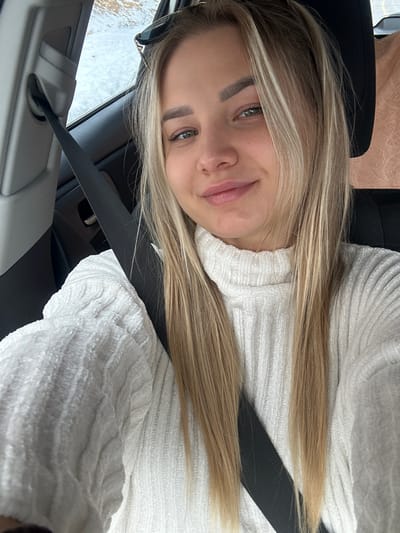 A young woman with blond hair is seated in a car wearing a seatbelt and a white sweater. The car appears to be moving on a rainy day. The background outside the car window shows a blurred image, suggesting motion. The image does not include any information connecting it to Donja Brezna, Montenegro.