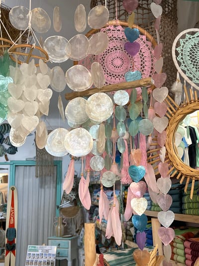 The image shows a close-up view of a display of decorative items, likely in a shop in Port de Pollença, Spain. The focus is on a variety of hanging ornaments, including numerous disc-shaped and heart-shaped pieces made from what appears to be mother-of-pearl or similar iridescent materials. These are strung together on threads and some also feature pink feathers and tassels. In the background, a circular woven mirror is visible, along with shelves stocked with colorful textiles. To the left, a vintage ski is propped up against a white curtained window, and a small desk with drawers is also present. The overall aesthetic suggests a bohemian or coastal theme, commonly found in tourist destinations like Port de Pollença. There are no people visible in the frame.
