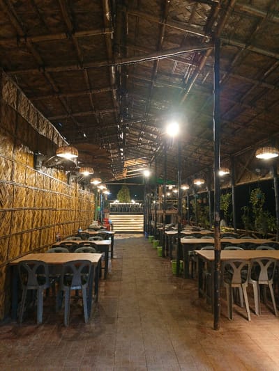 An indoor restaurant space with rows of dark wooden tables and chairs, extending into the distance. The structure is made of bamboo, creating a rustic and textured ambiance. Lights are hung above each table. The floor is brick and there is a brightly lit area in the distance, possibly an elevated stage. Green trash cans are visible next to each table.