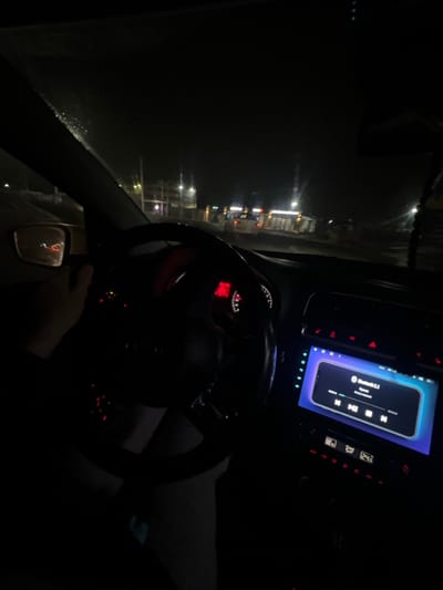 Inside a car at night, a steering wheel dominates the view with dashboard lights visible. To the left, a dark street scene unfolds through the window, dotted with faint streetlights. The car's entertainment system glows with a music player interface. A hand is visible near the gear stick. The overall scene is dim and shadowy, emphasizing the car's interior against the obscured urban backdrop.