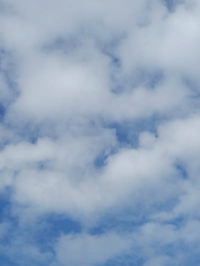 A sky filled with white, puffy clouds against a bright blue background is depicted in this vertical, full-frame, close-up shot. The image is underexposed, with the clouds appearing slightly blown out and the blue sky a muted tone. The scene is likely captured in Kyzyl Suu, Kyrgyzstan, given the geographic context provided. There are no visible people, subjects, objects, or actions in the image; it is a pure nature photograph of the sky. The environment is characterized by the atmospheric conditions of clouds and sky.