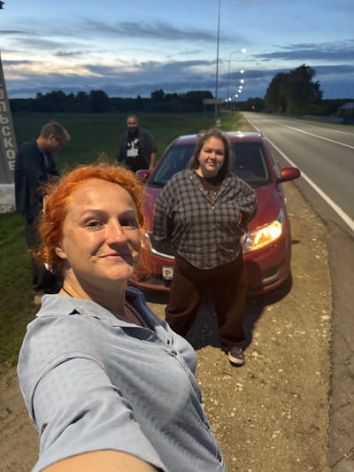 Four individuals are visible outdoors by a roadside at dusk. A woman with bright orange hair, wearing a light blue button-up shirt, is in the foreground, facing the camera and smiling. Behind her, a red car is parked on the shoulder of a road. To the left of the car and further back, a man with short brown hair and glasses, wearing a dark blue jacket, stands with his head down. Behind him, another man in a dark shirt with a graphic print is partially visible. To the right of the car and behind it, a woman with long brown hair is standing, wearing a checkered shirt and brown pants. The environment consists of a paved road with white lane markings, a gravel shoulder, a grassy field, and trees in the distance. Streetlights are visible along the road, and the sky is cloudy with a hint of sunset colors. A sign with Cyrillic text, including "ОЛЬСКОЕ", is partially visible on the left edge of the image, indicating the location is near Nikolskoye, Russia.