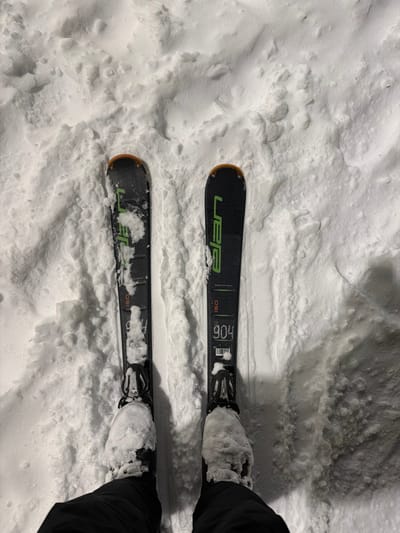 An overhead, first-person perspective captures an individual standing in deep, textured white snow in Makarovo, Russia. The person is wearing black ski pants and black ski boots, both of which are significantly covered with accumulated snow. Two black Elan brand skis are attached to the boots; these skis feature green branding and orange accents at their tips and tails. The right ski displays the markings "904" and "150", while the left ski shows "150". The bindings on both skis are visible and partially obscured by snow. The surrounding snow surface appears undisturbed, indicating a static position.