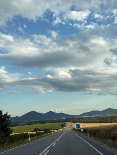 The image depicts a landscape view from a road in Luhyňa, Slovakia. A paved road with white lane markings extends into the distance, flanked by fields of green and golden crops. Rolling hills and dark, conical mountains form the backdrop under a sky filled with scattered white and gray clouds. Several blue road signs are visible along the side of the road. The scene appears to be an outdoor, rural environment during daylight. There are no people or animals visible in the image.