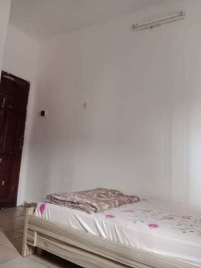 The image shows a simple bedroom in Kwale, Nigeria. The room is sparsely furnished with a bed in the center. The bed has a white sheet with a floral pattern and a blanket folded at the foot. The walls are painted white, and a fluorescent light fixture is mounted on the ceiling. A dark wooden door is visible on the left side of the frame. The flooring appears to be tiled. The scene is unremarkable and suggests a functional, no-frills living space.