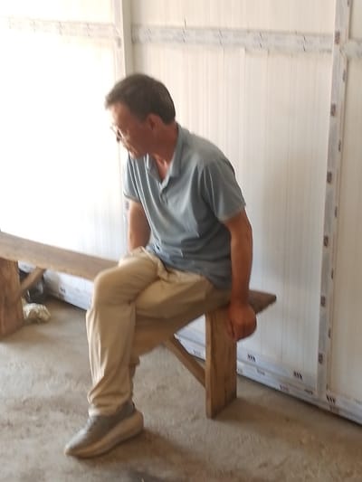 A middle-aged male is seated on a rustic wooden bench indoors in Olomoda, Nigeria. He wears a light blue short-sleeved polo shirt, khaki trousers, light-colored sneakers, and glasses. The man leans slightly forward, looking downwards and to his left, with his left leg crossed over his right. His right hand rests on his right knee. The background consists of a light-colored, vertically paneled wall with white framing elements, some featuring "WW" markings. The floor is concrete.