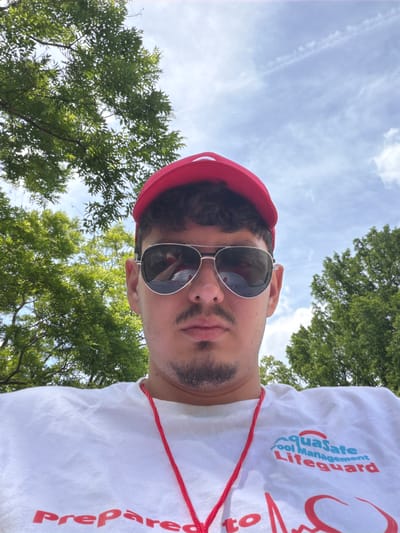 A man wearing a white shirt with the Aquasafe Pool Management Lifeguard logo and the word "Prepared" is shown in a selfie taken outdoors, possibly in Edison, United States of America. He is also wearing a red hat and sunglasses. Trees and a partly cloudy sky are visible in the background.