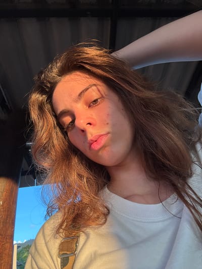 The image depicts a close-up, low-angle portrait of a young adult female, located in Ban Mae Ping, Thailand. She possesses medium-toned skin, brown eyes, and full, slightly parted lips. Her long, wavy brown hair, with reddish-brown highlights, is illuminated by direct golden-hour sunlight originating from the upper right. Multiple small blemishes or freckles are visible on her cheeks and nose. She is dressed in a white long-sleeved crew-neck top. A camouflage-patterned strap, likely from overalls or a bag, is visible over her left shoulder. Her right arm is raised, with the hand positioned out of frame, appearing to interact with her hair or head. The sunlight intensely brightens the right side of her face and hair, while the left side remains in shadow. The background features a dark, corrugated structure, possibly the underside of a roof or overhang, in the upper portion of the frame. A dark, vertical wooden pillar is visible on the far left. Below these structures, a clear blue sky, with hazy distant mountains, is visible in the lower-left section.