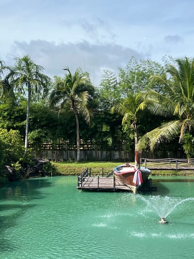 The image depicts a serene waterscape in Tambon Pa Kae Bo Hin, Thailand. The foreground is dominated by a vibrant, turquoise-colored body of water with ripples created by a fountain. Moored to a wooden pier in the middle ground is a traditional Thai longtail boat, adorned with colorful fabric. A wooden bridge stretches across the water to the right of the boat, with a simple wooden railing. The background features lush greenery, including tall palm trees and bamboo stalks, against a cloudy sky. The scene is tranquil and idyllic, suggesting a peaceful natural setting within the specified location.