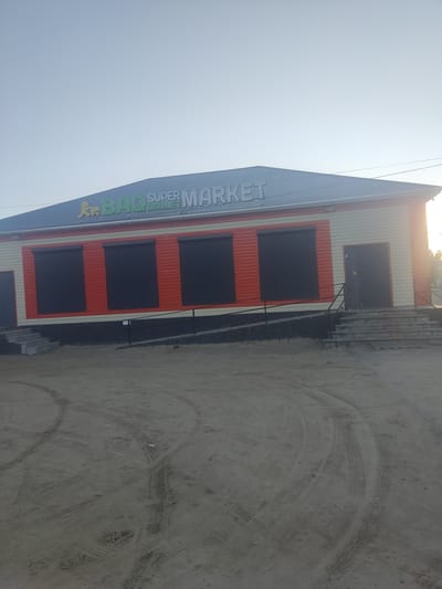 The image shows a low-angle, wide shot of a supermarket in the city of Aral, Kazakhstan. The supermarket has a prominent sign that reads "BAQ SUPERMARKET". The building has a light-colored facade with orange siding and black roll-up shutters on its windows. There is a dark-colored door on the right side of the building, accessible by a set of concrete stairs. The ground in front of the supermarket is dirt and shows tire tracks, suggesting a vehicle has recently been present. The sky is a clear, pale blue, with the sun low on the horizon, indicating it is either early morning or late afternoon.