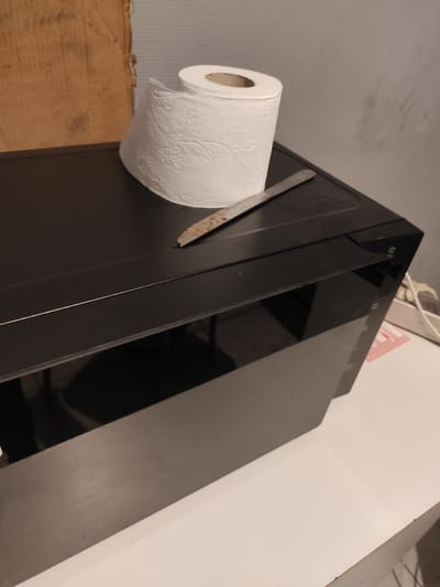 In Chaponost, France, on a white surface, rests a black, rectangular electronic device. On top of this device, a roll of toilet paper with a floral pattern is placed. Partially under the toilet paper, a small knife with a metallic blade, showing traces of rust and reddish residue, is positioned. To the left of the electronic device, a large, vertical, brown cardboard object stands. The scene is indoors, with a grey, textured wall in the background.