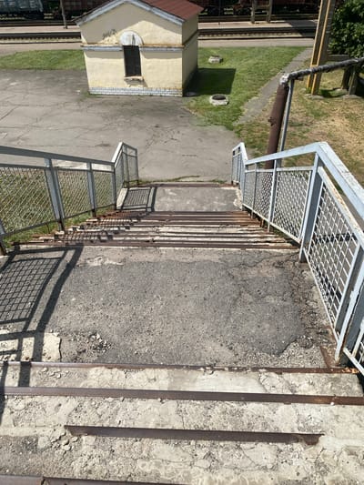 The image captures a weathered concrete staircase with metal railings, leading down from a slightly elevated area. The stairs are aged, with visible cracks and missing pieces of concrete, and reinforced with rusty metal strips. The railings are made of metal mesh and painted a faded grey.

In the background, a small, light yellow building with a dark-paned window is visible, situated near a grassy area. Adjacent to the grassy area are railway tracks with what appear to be train cars. The ground surrounding the stairs is a mixture of cracked asphalt and gravel. The lighting suggests it is daytime, with strong shadows cast by the railings and the nearby building.

This scene is located at the Romodan railway station in Romodan, Ukraine.