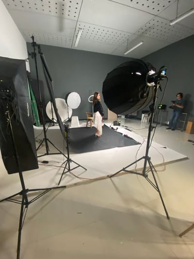 Inside a photo studio, a woman in a white dress poses on a black platform. Softboxes and lighting equipment surround her. A photographer stands in the background, capturing the scene. The studio has gray walls with ventilation holes and fluorescent lights. Large tripods and diffusion panels complete the setup, creating a controlled lighting environment for the photoshoot.