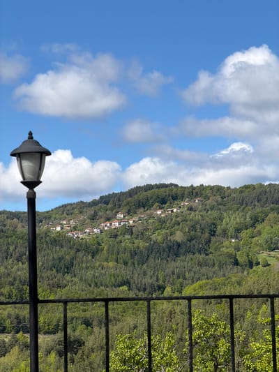 The image shows a view from a balcony in Кирково, Bulgaria. A black metal railing is partially visible on the right side. In the background, a green hillside is dotted with houses. A lamp post with a white lamp stands out against a blue sky with white clouds.
