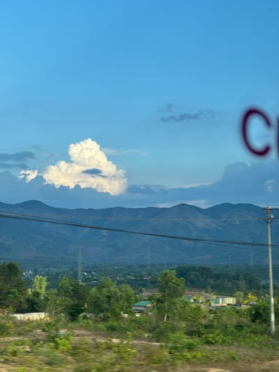 The image shows a landscape near Ia Khươl, Vietnam. In the foreground, there are trees and greenery, blending with a brownish, coarse terrain. Midground features rolling hills or mountains in a muted blue hue with electricity pylons cutting through the landscape. The sky is predominantly blue, punctuated by fluffy, white clouds. The presence of a blurred, red circular shape suggests the photo was taken through a window.