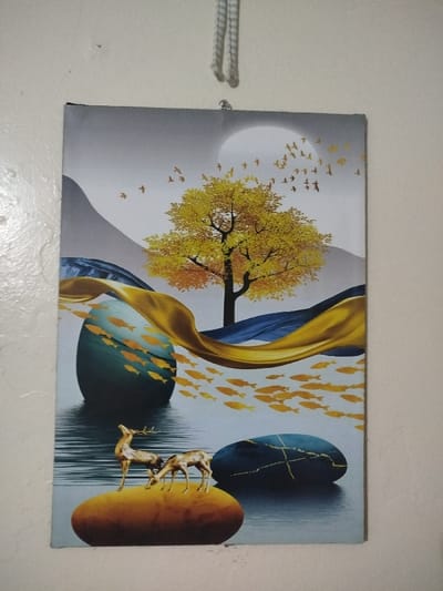 An art piece hangs on a wall, featuring a stylized landscape. A golden tree stands under a bright moon, with a flock of birds flying overhead. Golden fish swim around abstract shapes, including large rocks and a flowing golden ribbon. Two deer stand on a prominent orange rock. The color palette is muted, with blues, golds, and grays dominating the scene.
