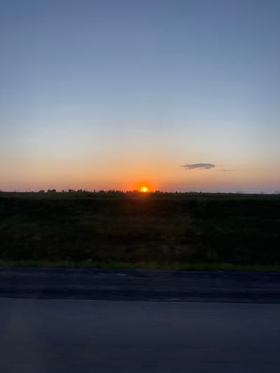 The image shows a sunset at Kamennoye, Russia. The sun is setting behind a treeline, casting an orange glow on the sky. To the left of the treeline, the sky is a light blue. There is a small cloud above the sun. A dark surface, possibly a road or wall, is present on the right of the image.