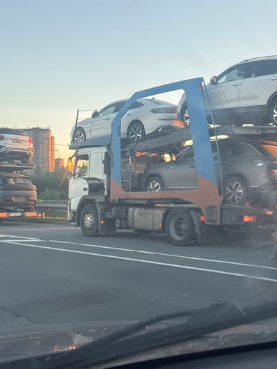 An auto transport truck is shown on a highway with a golden sky suggesting either sunrise or sunset. The truck is carrying multiple cars on its upper decks. One car, a white sedan, is clearly visible on the top deck. Below it, a dark colored SUV is visible on a lower deck. In the background, a cluster of tall buildings, likely apartment blocks, are illuminated by the setting or rising sun. The truck appears to be in transit, likely on a road in or near Poroshkino, Russia, given the architectural style of the buildings. The perspective of the image is from inside another vehicle, looking out at the truck.