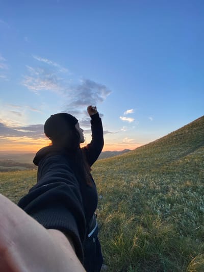 A person stands on a grassy hill, looking towards a sunset over a valley. The person is wearing a black beanie and a black hoodie, and their right arm is raised. The sky is a blend of blue and orange, with scattered clouds. The hill is covered in green grass and some low-lying vegetation. In the distance, mountains are visible. The overall scene is serene and natural. The location is specified as Абзаново, Russia.