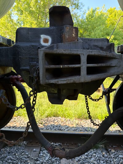 The image depicts a close-up view of a train's coupling mechanism, likely in an outdoor setting. The primary subject is a heavy, dark metal coupling device, featuring a robust, box-like structure with internal crossbars. Attached to this coupling is a thick, dark hose, secured by metal clamps. A rusty metal chain is also visible, connected to the coupling and extending out of frame. The background shows blurred green foliage, suggesting trees, and a clear blue sky, indicating daylight. The ground beneath appears to be covered in gravel and railway ties, confirming the location is near train tracks. There are no people visible in the image, and the overall impression is one of industrial machinery in a natural environment. The image is oriented vertically. There is a small, irregular white marking with a reddish-brown center on the side of the coupling mechanism. No visible text is present in the image.