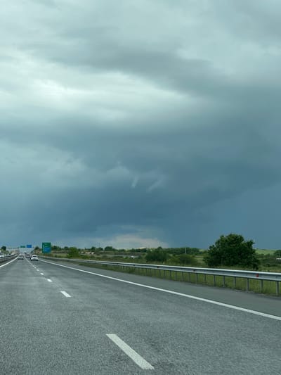 The image shows a highway in the city of Svoboda, Bulgaria. The road surface is gray asphalt, with white lane markings. A metal guardrail runs along the edge of the road. Beyond the guardrail, there is greenery and trees. A line of vehicles is visible in the distance. The sky is overcast with dark clouds.