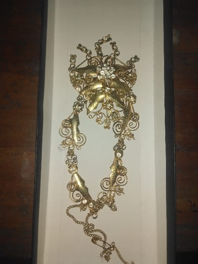 An intricate piece of gold-toned jewelry is displayed vertically within a white-lined rectangular container, positioned against a dark wooden surface. The jewelry features a large central cluster of clear, faceted stones, flanked by numerous smaller stones and stylized gold leaves. Ornate scrollwork and additional elements extend downwards, forming a chain-like structure to which a small metal safety pin is affixed. There are no individuals, actions, or interactions depicted. This scene is located in the city of Gerasimovskoye, Kazakhstan.
