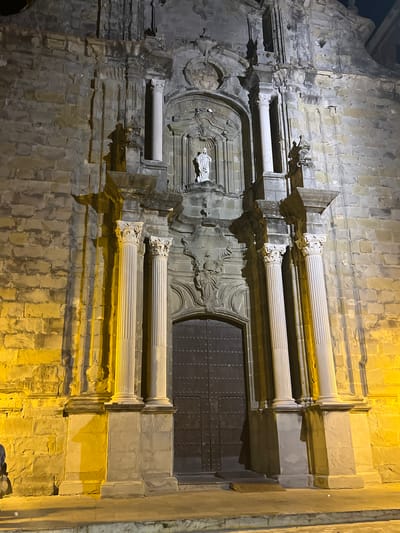 This image captures the facade of a historic stone church at night, illuminated by artificial yellow lighting. The architecture features classical elements such as fluted columns, ornate carvings, and a statue of a robed figure positioned above a large, dark wooden door. The stone blocks of the facade are weathered, and some show signs of age and repair. Shadows cast by the architectural details and the lighting create a dramatic effect. The ground in front of the church is paved, with a few small debris items visible. The overall scene suggests an ancient religious structure in a European city. The church is located in Tarifa, Spain.