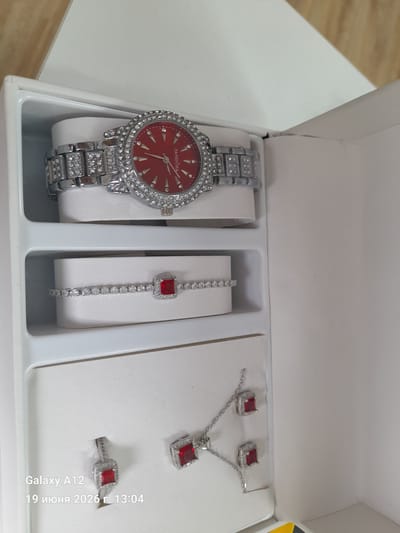 The image shows a jewelry set in a white box. The set includes a watch with a red face and a diamond-encrusted band, a bracelet with a red stone and small diamonds, and a necklace with a red stone and a chain. The box is open, and the jewelry is displayed on white velvet pads. The background is a plain white surface, with a hint of wooden texture at the top. There is no indication of the location in the city of Kara-Köl City, Kyrgyzstan.