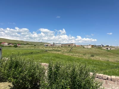 The image depicts a rural landscape under a bright blue sky dotted with fluffy white clouds. In the foreground, lush green bushes and a low stone wall create a natural frame. Beyond this, rolling hills covered in vibrant green grass stretch towards the horizon. Several scattered buildings, including what appear to be farm structures and houses with red roofs, are visible on the hillsides. Some of these buildings are under construction, with scaffolding and exposed frameworks. The scene suggests a clear, sunny day, likely in the late morning or early afternoon given the bright sunlight and distinct shadows. The overall impression is one of a peaceful, undeveloped rural area in Armenia, possibly on the outskirts of a village or town. No people are visible in the image, and the focus is on the natural landscape and the man-made structures within it. There is some visible text on a yellow object in the distance, but it is too small and blurry to read.