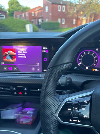 The image shows the interior of a car, specifically the driver's side, with a focus on the steering wheel, dashboard, and infotainment system. The infotainment screen displays the song "Plain Jane" by ASAP Ferg from the album "Still Striving." The digital instrument cluster behind the steering wheel shows the car is in "D" drive mode. The steering wheel has cruise control buttons. A residential area with brick buildings and trees is visible through the windshield, suggesting the car is parked or driving in a suburban neighborhood. A box is visible in the center console of the car.