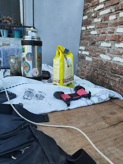 A wooden table is topped with a white towel with a subtle pattern. A metal thermos with stickers, a glass jar, and a yellow bag of "Playadito" yerba mate sit atop the towel. A black bag with straps and a white phone charging cable are in the foreground. A red and black object resembling a controller extension is also on the towel. The background features a window with plants and a light blue wall transitioning into a brick wall.